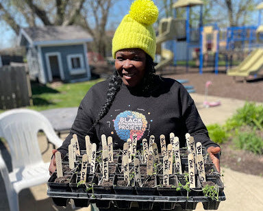 Person holding a tray of seedlings in an outdoor setting with playground equipment in the background.
